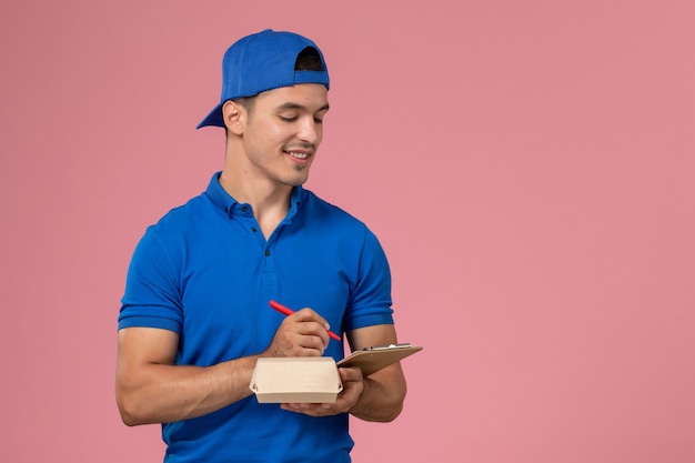 Front view young male courier in blue uniform cape holding little delivery food package and notepad writing notes on the light-pink wall