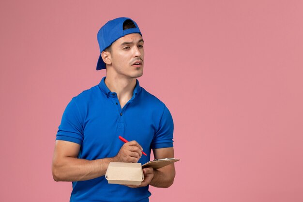 淡いピンクの壁にメモを書く小さな配達食品パッケージとメモ帳を保持している青い制服ケープの正面図若い男性の宅配便