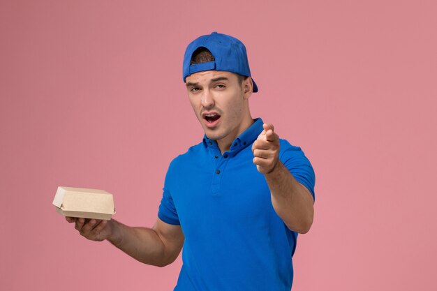 Front view young male courier in blue uniform cape holding little delivery food package on the pink wall