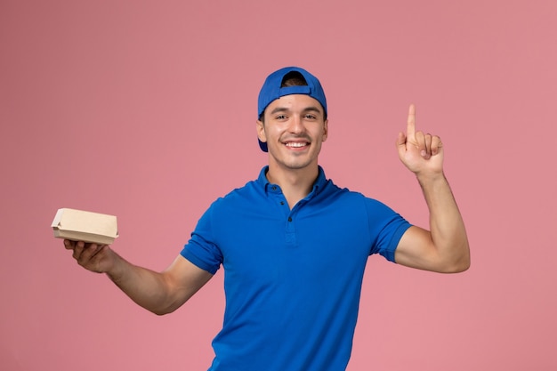 Front view young male courier in blue uniform cape holding little delivery food package on the pink wall