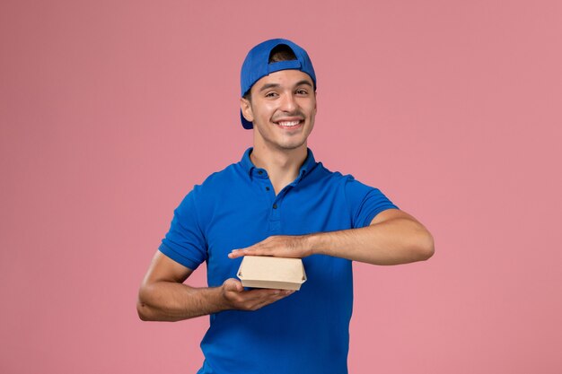 Front view young male courier in blue uniform cape holding little delivery food package smiling on the pink wall