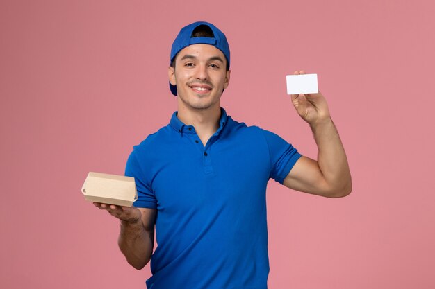 Front view young male courier in blue uniform cape holding little delivery food package and white card on light pink wall