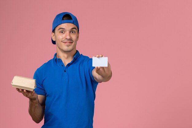 Вид спереди молодой курьер-мужчина в синей форменной накидке с небольшим пакетом еды для доставки и белой карточкой на светло-розовой стене