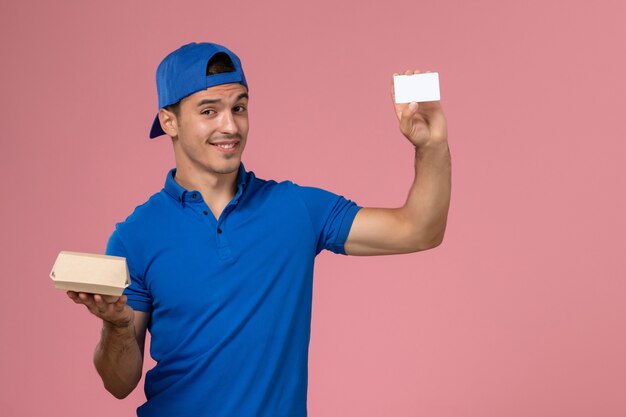 Front view young male courier in blue uniform cape holding little delivery food package and white card on light pink wall