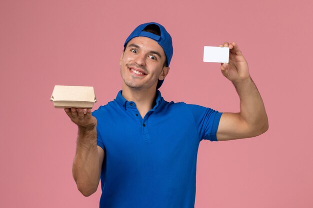 Вид спереди молодой курьер-мужчина в синей форменной накидке с небольшим пакетом еды для доставки и белой карточкой на светло-розовой стене