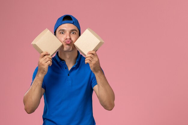 Вид спереди молодой курьер-мужчина в синей форменной накидке с небольшими пакетами еды для доставки на светло-розовой стене