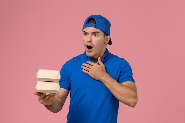 Вид спереди молодой курьер-мужчина в синей форменной накидке с небольшими пакетами еды для доставки на розовой стене