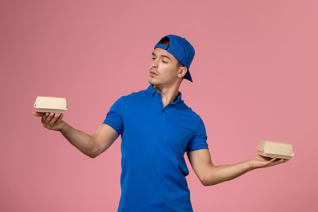 Front view young male courier in blue uniform cape holding little delivery food packages on the pink wall