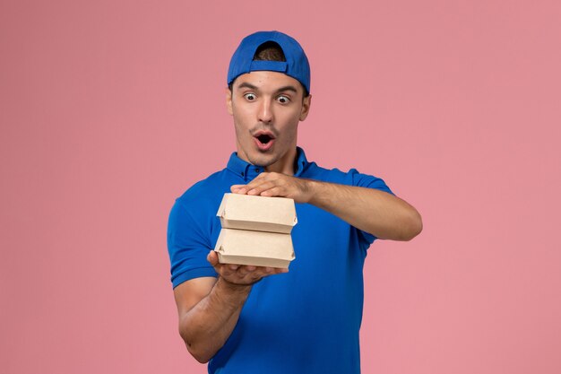 Front view young male courier in blue uniform cape holding little delivery food packages on the pink wall