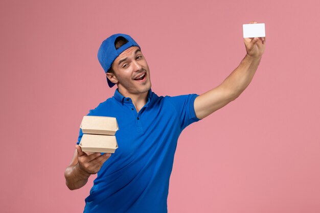 淡いピンクの壁にカードと小さな配達食品パッケージを保持している青い制服の岬の正面図若い男性の宅配便