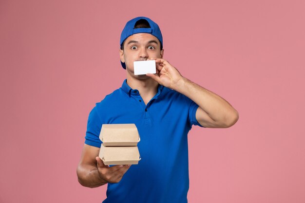 Front view young male courier in blue uniform cape holding little delivery food packages with card on light pink wall