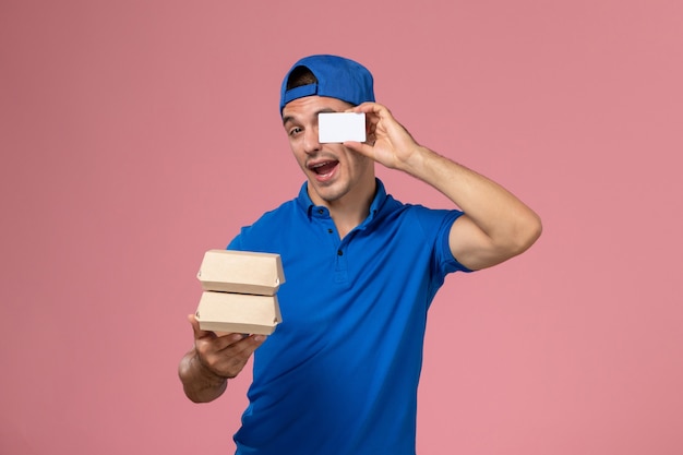 Front view young male courier in blue uniform cape holding little delivery food packages with card on light pink wall