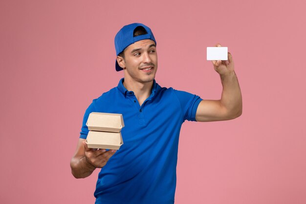 Front view young male courier in blue uniform cape holding little delivery food packages with card on light pink wall