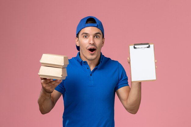 Front view young male courier in blue uniform cape holding little delivery food packages with notepad on light pink wall