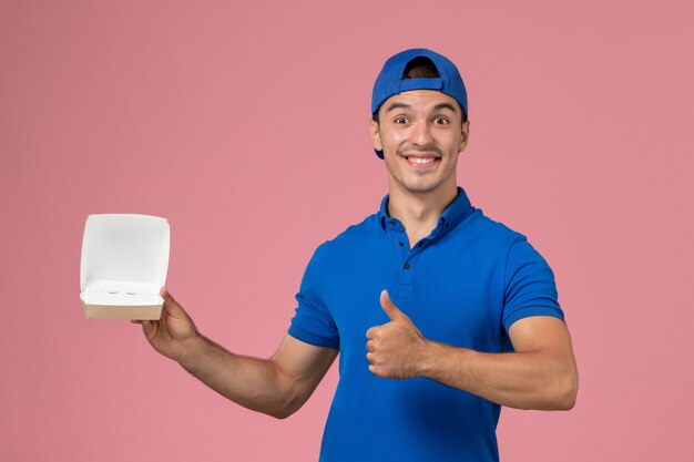 Front view young male courier in blue uniform cape holding little empty delivery food package on the pink wall