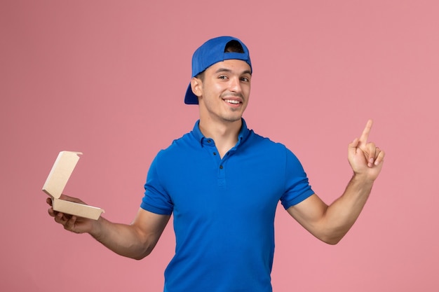 ピンクの壁に笑みを浮かべて少し空の配達食品パッケージを保持している青い制服ケープの正面図若い男性宅配便