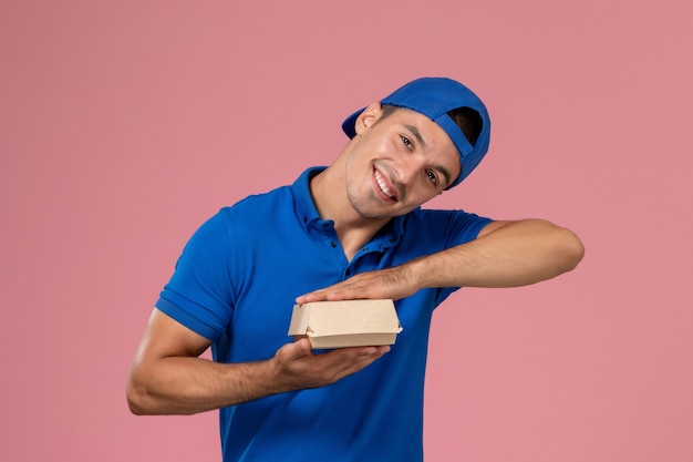Front view young male courier in blue uniform cape holding little empty delivery food package with smile on the pink wall