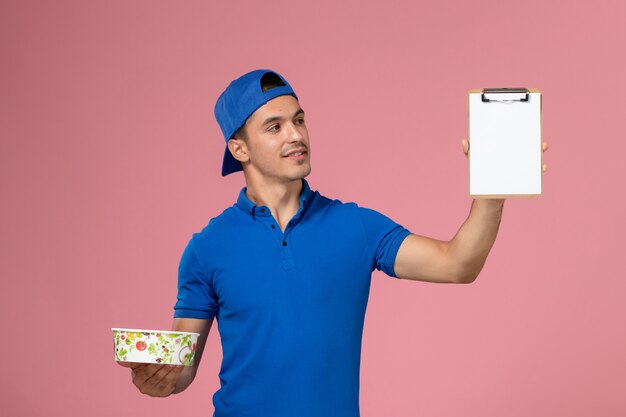 Front view young male courier in blue uniform cape holding notepad and round delivery bowl on the light pink wall