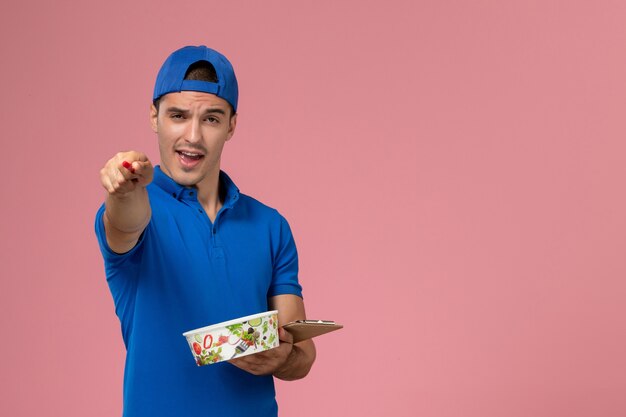 Front view young male courier in blue uniform cape holding notepad with pen and round delivery bowl on pink wall