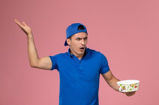 Вид спереди молодой курьер-мужчина в синей форменной накидке держит круглую миску для доставки на светло-розовой стене