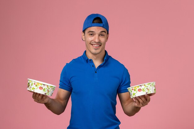 Front view young male courier in blue uniform cape holding round delivery bowls on light pink wall