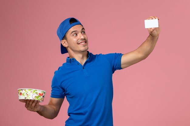 Front view young male courier in blue uniform cape holding white card and round delivery bowl on light pink wall