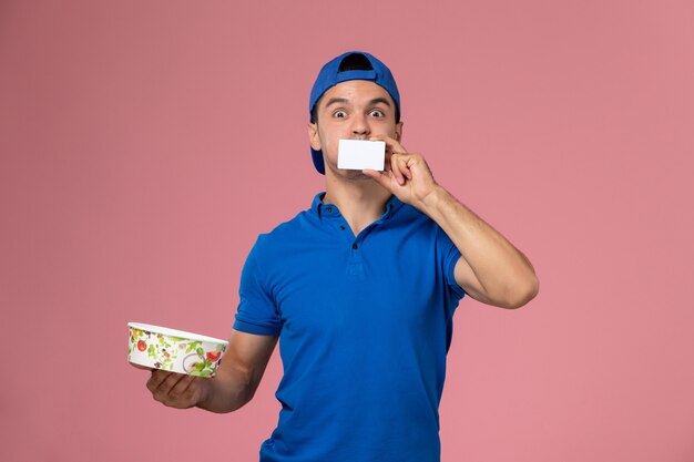 Front view young male courier in blue uniform cape holding white card and round delivery bowl on light pink wall