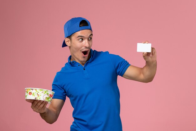 Вид спереди молодой курьер-мужчина в синей форме, держащий белую карточку и круглую миску для доставки на светло-розовой стене
