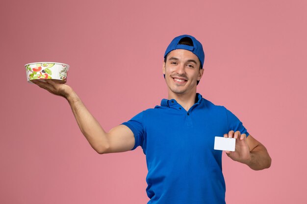 Front view young male courier in blue uniform cape holding white card and round delivery bowl smiling on light pink wall