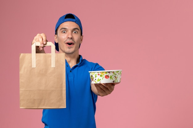 Front view young male courier in blue uniform and cape with delivery package and bowl on his hands on the pink wall