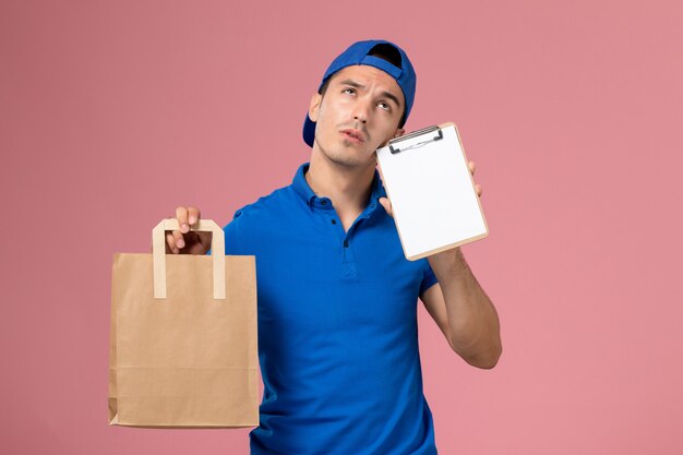 Front view young male courier in blue uniform and cape with delivery package and notepad on his hands on pink wall
