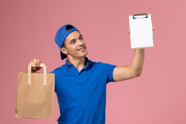 Front view young male courier in blue uniform and cape with delivery package and notepad on his hands on pink wall