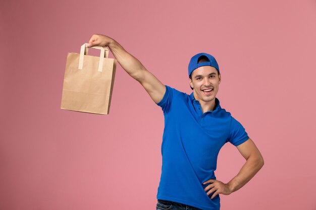 Front view young male courier in blue uniform and cape with delivery paper package on his hands on pink wall