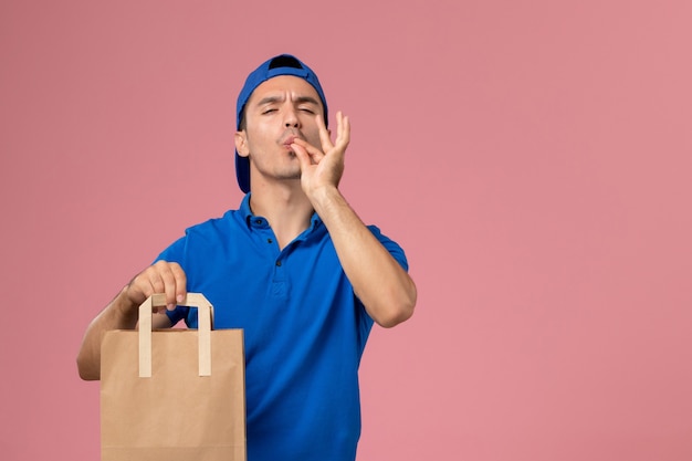 Front view young male courier in blue uniform and cape with paper delivery package on his hands on pink wall