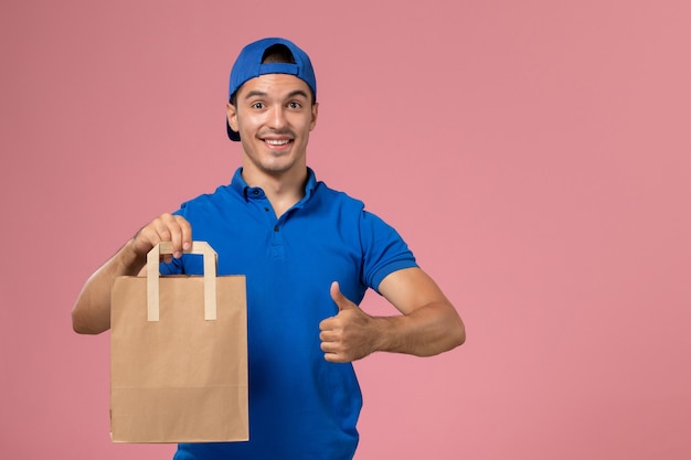 Front view young male courier in blue uniform and cape with paper delivery package on his hands on pink wall