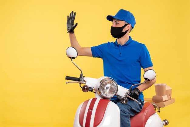 Front view young male courier in blue uniform on yellow background covid- job pandemic service virus bike work