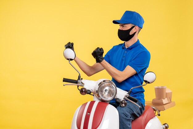 Front view young male courier in blue uniform on the yellow background covid- pandemic delivery service job virus bike work