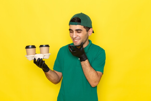 Front view young male courier in green shirt green cap holding coffee cups and talking on phone with laugh on yellow