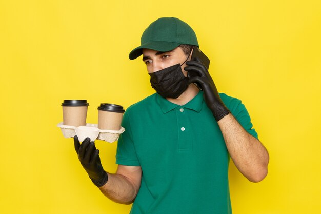 Front view young male courier in green shirt green cap holding coffee cups and talking on phone on yellow