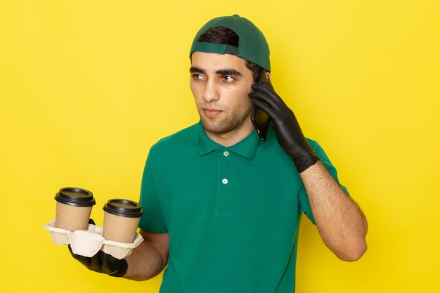 Front view young male courier in green shirt green cap holding coffee cups and talking on phone on yellow