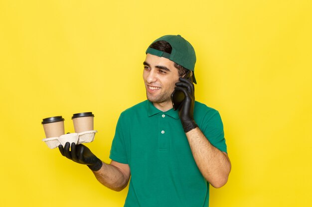 Front view young male courier in green shirt green cap holding coffee cups and talking on phone on yellow