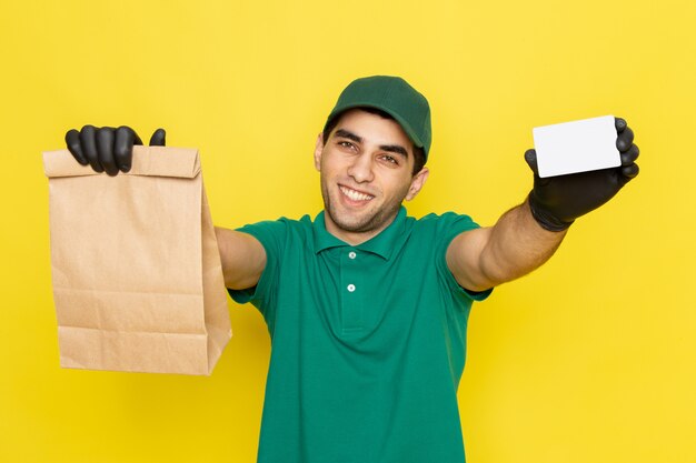 Front view young male courier in green shirt green cap holding delivery package and white card on yellow