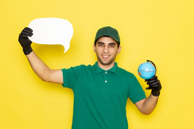 Front view young male courier in green shirt green cap holding little globe and white sign with black gloves on yellow