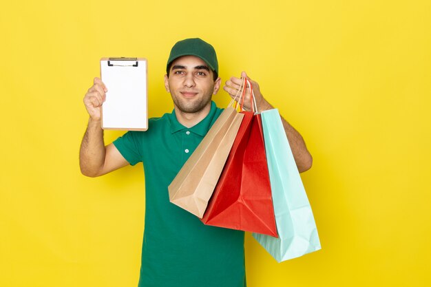 黄色のショッピングパッケージとメモ帳を保持している緑のシャツグリーンキャップの正面の若い男性宅配便