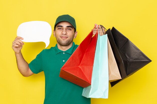 Front view young male courier in green shirt green cap holding shopping packages and white sign on yellow