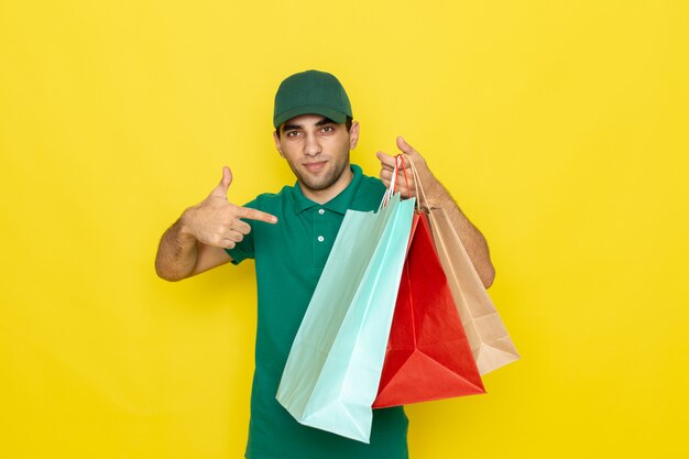 Front view young male courier in green shirt green cap holding shopping packages with smile on his face on yellow