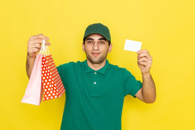 黄色の白いカードとショッピングパッケージを保持している緑のシャツグリーンキャップの正面の若い男性宅配便