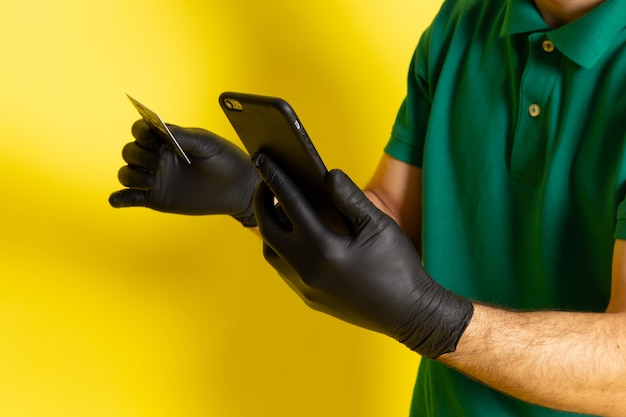 Front view young male courier in green shirt green cap holding white card using his smartphone on yellow