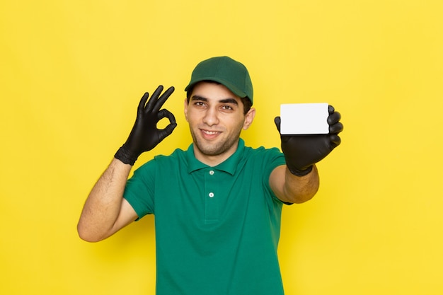 Front view young male courier in green shirt green cap holding white card on yellow