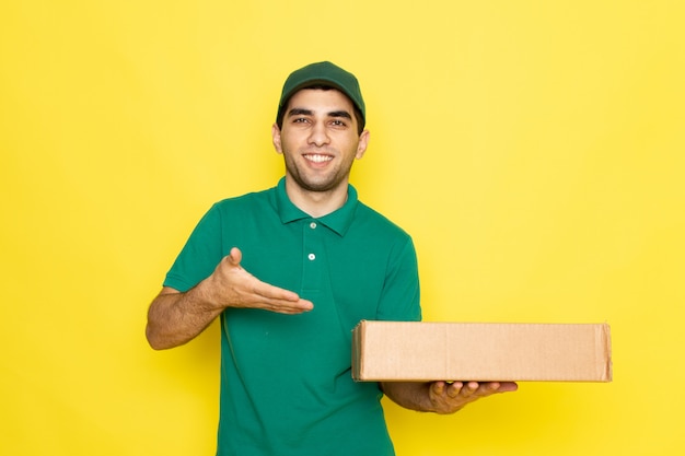 Front view young male courier in green shirt green cap smiling and holding delivery box pointing into delivery box on the yellow background delivering service color